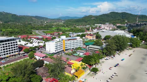 Stunning-aerial-view-of-a-bustling-tropical-beach-lined-with-resort-buildings,-scenic-hills,-and-calm-turquoise-waters-in-Malaysia