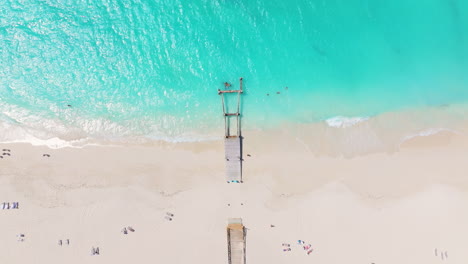 Stunning-aerial-4K-view-of-Grace-Bay-Beach-featuring-a-central-pier,-glistening-turquoise-waters-with-gentle-waves,-and-sunbathers-on-the-pristine-white-sand