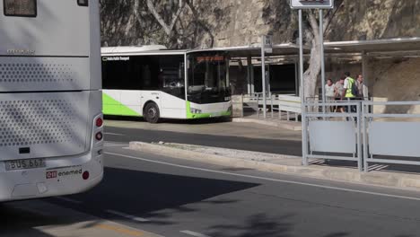 At-the-bus-station-in-Valletta,-Malta,-buses-are-stationed-to-pick-up-travelers-while-others-pass-by,-illustrating-the-bustling-transportation-hub-and-commuter-life
