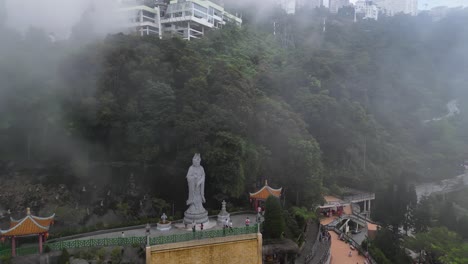 Aerial-View-Foggy-day-at-a-Malaysian-temple-with-a-large-statue-surrounded-by-lush-greenery-and-distant-buildings