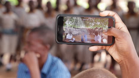 Ein-Schwarzer-Afrikanischer-Lehrer-Hält-Ein-Modernes-Smartphone-In-Der-Hand-Und-Filmt-Dabei-Eine-Gruppe-Von-Schülerinnen-In-Uniform,-Die-In-Einer-Abgelegenen-Dorfschule-Ein-Traditionelles-Indigenes-Ritual-Tanzend-Und-Singend-Durchführen