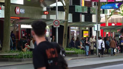 Commuters-waiting-at-the-Bourke-Street-Mall-and-Swanston-St-tram-stop-in-the-bustling-downtown-Melbourne-city,-with-pedestrians-crossing