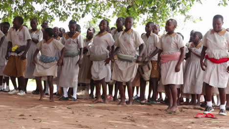 Un-Grupo-De-Estudiantes-Africanos-Vestidos-Con-Uniforme-Blanco-Realizan-Juntos-Una-Danza-Ritual-En-Una-Aldea-Remota-Tocando-Tambores,-Aplaudiendo-Y-Cantando.