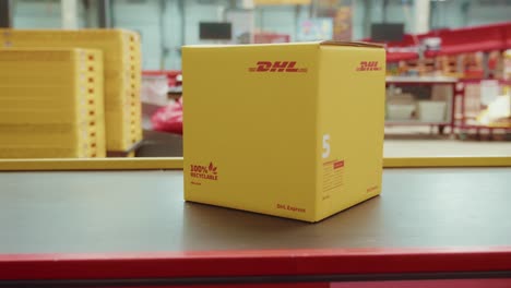 Caja-De-Cartón-Amarilla-Con-El-Logotipo-De-DHL-En-La-Línea-De-Cinta-Transportadora-Dentro-Del-Almacén