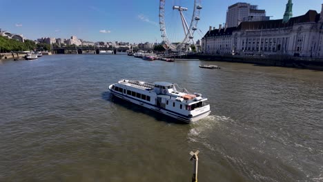 A-white-riverboat-cruises-through-the-Thames-River-in-London,-passing-by-the-iconic-London-Eye-Ferris-wheel-under-a-clear-blue-sky,-with-visible-historic-buildings-in-the-background