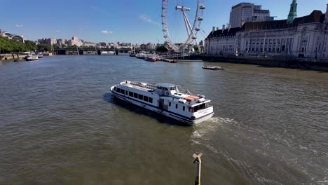 Ein-Weißes-Flussboot-Fährt-Auf-Der-Themse-In-London-Vorbei-Am-Berühmten-Riesenrad-London-Eye-Unter-Einem-Klaren-Blauen-Himmel,-Im-Hintergrund-Sind-Historische-Gebäude-Zu-Sehen