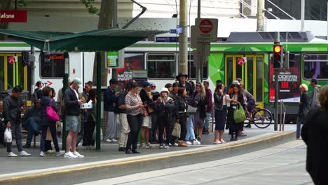 Commuters-waiting-at-the-Swanston-St-and-Bourke-St-tram-stop-in-the-bustling-downtown-Melbourne-city,-with-pedestrians-crossing