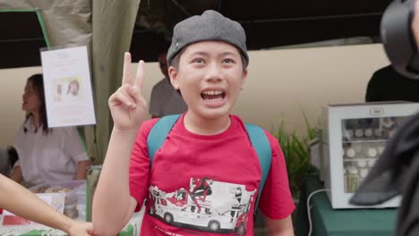 Young-boy-with-autism-at-an-outdoor-event,-smiling-and-giving-a-peace-sign-while-a-woman-stands-nearby