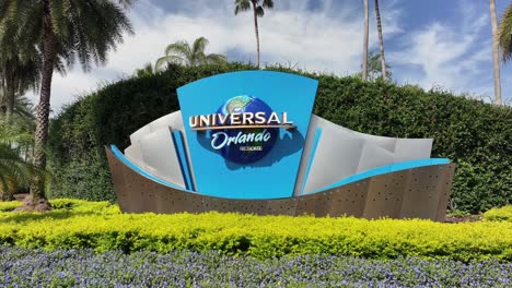 Universal-Orlando-Resort-sign-surrounded-by-lush-greenery,-colorful-flowers,-and-palm-trees,-set-against-a-partly-cloudy-blue-sky