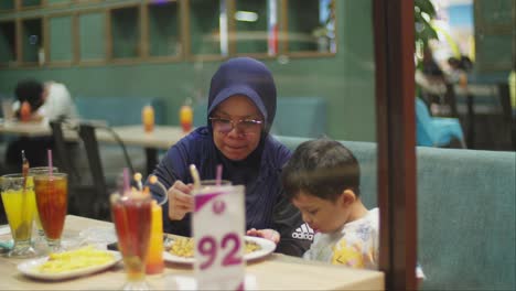 Madre-Indonesia-Con-Hijab-Alimentando-A-Su-Hijo-En-Un-Restaurante