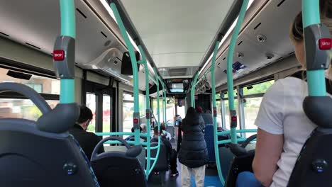 Interior-of-a-modern-city-bus-with-teal-handrails,-showing-passengers-seated-and-standing-with-focus-on-the-back-seats-and-displays-above