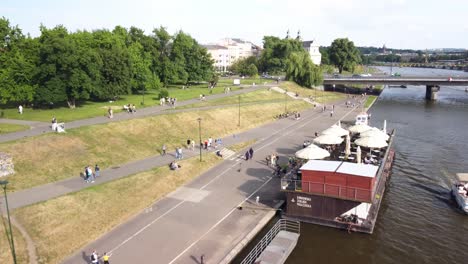 People-walking-and-relaxing-by-the-water,-moored-boats,-and-green-spaces-along-the-riverbank-in-Krakow,-Poland