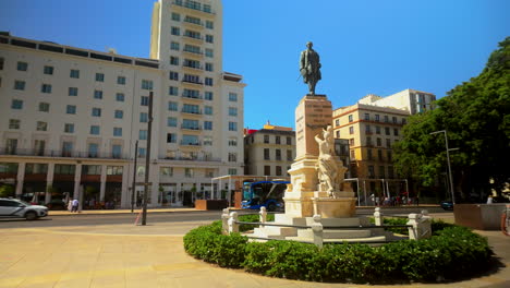 Día-Soleado-En-El-Monumento-Al-Marqués-De-Larios-En-Málaga,-Con-Edificios-Históricos-Y-Flores-Vibrantes-Cerca