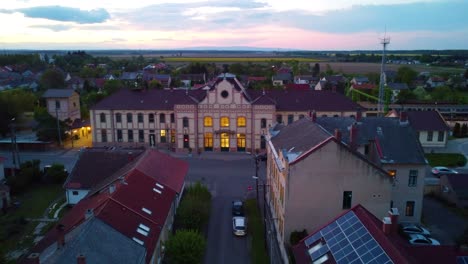 Eine-Drohnenaufnahme-Nähert-Sich-Dem-Historischen-Bahnhof-Von-Körmend,-Der-Von-Lichtern-Beleuchtet-Wird,-Während-Die-Sonne-über-Der-Kleinen-Europäischen-Stadt-Untergeht