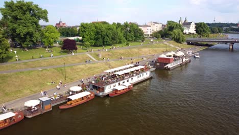 moored-boats,-waterfront-cafes,-and-people-enjoying-in-Wisla-River-promenade-in-Krakow