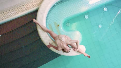 Flexible-European-female-gymnast-does-split-on-pool-side,-top-down-aerial-view