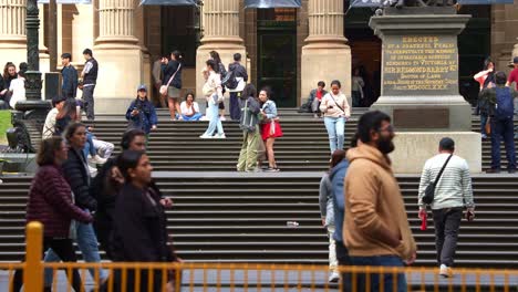 The-forecourt-staircases-of-State-Library-Victoria,-locals-and-visitors-hang-out-on-the-lawn-space-in-downtown-Melbourne's-bustling-Central-Business-District,-with-pedestrians-strolling-by