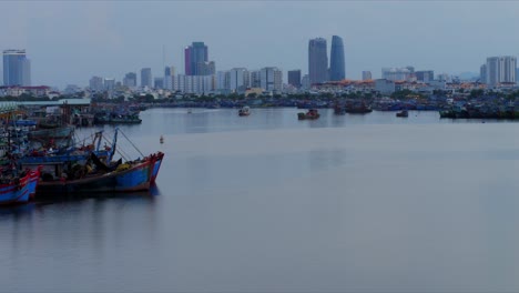 Timelapse-De-Día-A-Noche-En-El-Antiguo-Puerto-De-Da-Nang-Con-Un-Tradicional-Barco-De-Madera-Y-El-Horizonte-Urbano-De-La-Ciudad-Moderna-A-Distancia