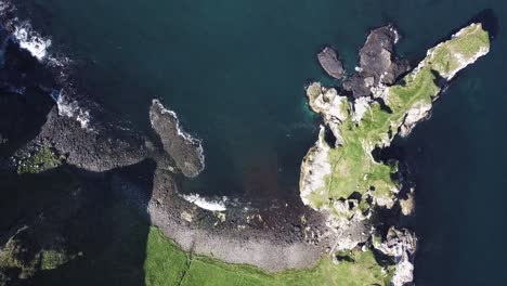 Top-down-drone-view-of-Casterly-Rock-Bay,-Ireland-with-waves-crashing-on-pebbles-beach-and-lush-green-mountains