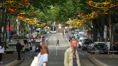 A-vibrant-Melbourne-street-scene-with-trams-glide,-pedestrians-stroll,-and-festive-decorations-creating-a-bustling-urban-atmosphere-in-the-central-business-district,-captured-in-slow-motion-shot