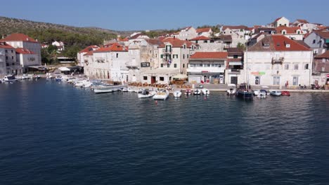 Croatian-flag-fluttering-above-boats-moored-in-harbor-of-medieval-Milna-town,-Drone-dolly-out