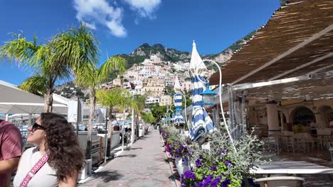 Gente-Caminando-Por-El-Paseo-Marítimo-De-La-Playa-De-Positano-Con-Hermosas-Casas-Costeras-Al-Fondo,-Costa-De-Amalfi