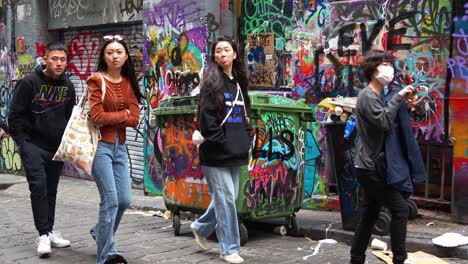 Tourists-visiting-Hosier-Lane-in-Melbourne,-a-cobblestone-laneway-featuring-a-vibrant-array-of-art-murals-and-graffiti-on-the-exterior-walls-of-buildings-with-overflowing-rubbish-from-the-wheelie-bins