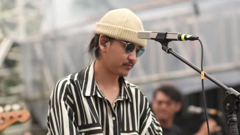 Asian-Stylish-Man-Musician-Performing-Live-On-Stage-With-Music-Band-During-Outdoor-Concert