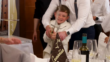 Un-Niño-Rompiendo-Una-Torre-De-Chocolate-Con-Una-Espada-Con-La-Ayuda-De-Su-Padre-Sobre-Su-Hombro-Durante-Una-Primera-Comunión.