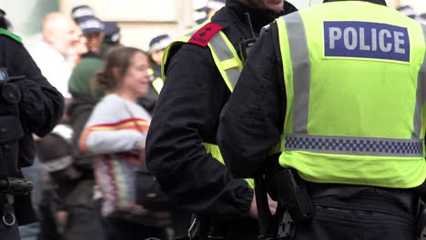 A-Metropolitan-Territorial-Support-Group-police-sergeant-and-inspector-stand-and-talk-as-people-walk-by-and-a-unit-of-officers-wait-for-orders-in-the-background-during-a-public-order-event