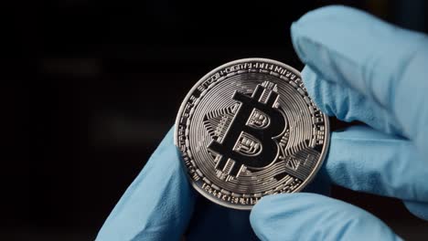 Hand-With-Blue-Gloves-Holding-Bitcoin-Crypto-Currency-Coin-Against-Black-Background,-Close-Up