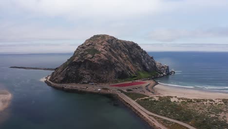 Wide-aerial-descending-shot-over-the-narrow-causeway-leading-to-Morro-Rock-in-Morro-Bay,-California