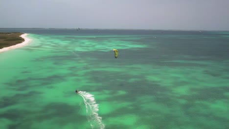 Kitesurfer-Mit-Gelbem-Drachen-Gleitet-über-Türkisfarbenes-Wasser-In-Der-Nähe-Einer-Weißen-Yacht,-Luftaufnahme