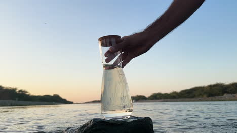 Hand-placing-a-caraf-with-fresh-water-on-a-rock-in-middle-of-a-river,-during-sunset