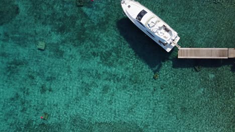 Eine-Weiße-Yacht,-Die-An-Einem-Holzsteg-Im-Klaren-Blauen-Strandwasser-Festgemacht-Ist,-Drohne-Von-Oben-Nach-Unten