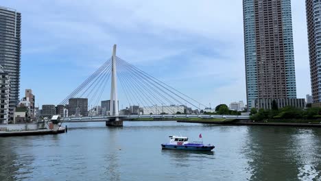 Boat-sails-on-river-with-cityscape-and-modern-bridge-in-the-background-under-a-clear-sky