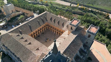 Vuelo-Ascendente-En-El-Monasterio-De-Uclés-Visualizando-El-Claustro-Con-Sus-Arcos,-El-Tejado-De-La-Iglesia-Con-Sus-Torres-De-Entrada-Y-La-Cabecera-De-Otra-Torre-Rematada-En-Punta-Oscura