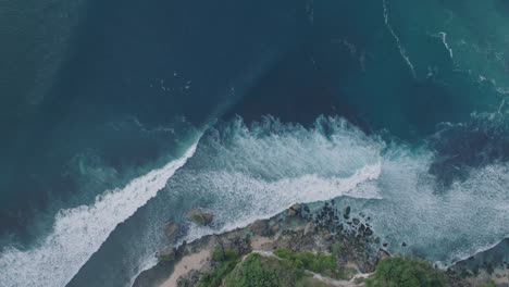 Drone-De-Arriba-Hacia-Abajo-De-Surfistas-No-Identificables-Al-Atardecer-Con-Agua-Azul-Profunda-Y-Dorada-Y-Costa-En-Padang-Padang,-Bali,-Uluwatu-Indonesia