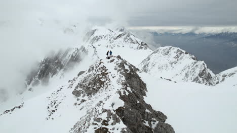 Revealing-Drone-Shot-of-Couple-Standing-on-Top-of-the-Rock-With-Amazing-View-of-Snow-Capped-Peaks