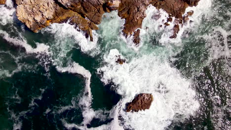 Top-down-aerial-of-ocean-waves-surging-against-jagged-rocky-coastline