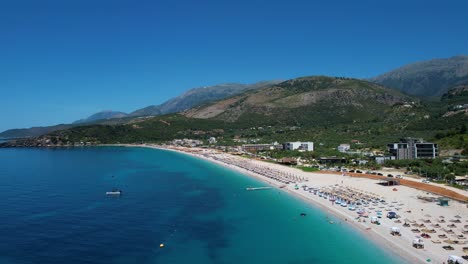 Beautiful-Coastline-with-Blue-Water,-White-Sand-Beach,-and-Green-Hills-in-Livadh-Bay,-Full-of-Summer-Holidaymakers-in-Umbrellas-and-Sunbeds