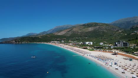 Hermosa-Costa-Con-Agua-Azul,-Playa-De-Arena-Blanca-Y-Colinas-Verdes-En-La-Bahía-De-Livadh,-Llena-De-Turistas-De-Verano-Con-Sombrillas-Y-Tumbonas