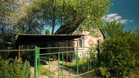 Aufnahmen-Eines-Sommerhauses-Aus-Backstein-Mit-Garten-Und-Einem-Grünen-Eisentor,-Das-Im-Frühling-Im-Wind-Schwankt