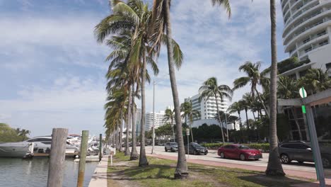 Scenic-Miami-Beach-road-and-waterway-lined-with-palm-trees,-luxury-yachts,-and-modern-high-rise-buildings-under-a-clear-sky