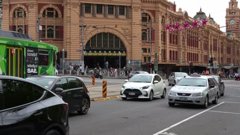 Tram-stops-at-the-traffic-light,-vehicles-traffic-on-Swanston-and-Flinders-streets-with-iconic-Flinders-Street-Station-entrance-as-backdrop,-creating-a-vibrant-transit-hub-for-commuters-and-tourists