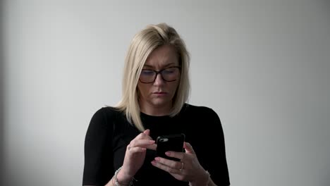 Blonde-woman-in-black-attire-with-glasses-sitting-in-an-office,-intently-using-her-smartphone