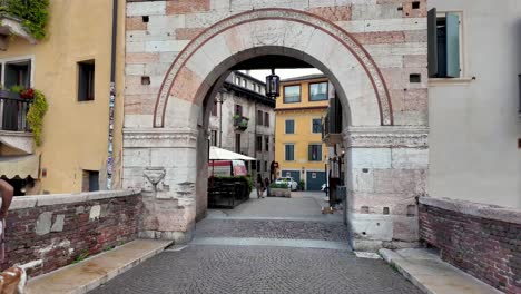 Walking-over-the-bridge-Ponte-Pietra,-through-arched-stone-gates,-over-small-square-and-streets-with-historical-architecture,-Verona-Italy