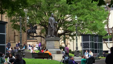 Landmark-statue-of-Sir-Redmond-Barry-to-commemorate-the-founder-of-State-Library-Victoria-located-on-the-forecourt,-people-hanging-out-and-relaxing-on-the-lawn,-a-lively-urban-life-in-Melbourne-city