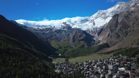 Sunny-stunning-Saastal-Saas-Fee-village-town-Switzerland-aerial-drone-beautiful-Fall-Autumn-Swiss-Alps-mountain-peaks-glacier-surrounding-city-buildings-bridge-Zermatt-The-Matterhorn-backwards