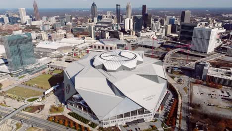 Drone-shot-of-Mercedes-Benz-Stadium-surrounded-by-office-buildings-in-autumn-afternoon-with-Downtown-Atlanta-skyscrapers-in-the-background